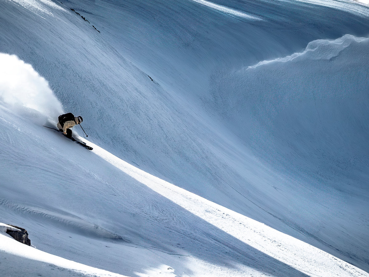 Skier descending a steep snow-covered mountainside, carving through deep powder in alpine terrain.