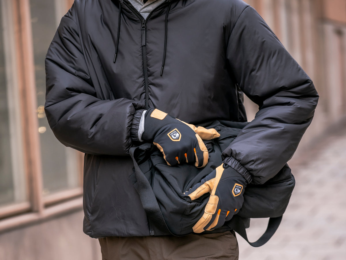 Black and beige men's gloves with leather details, worn with a black down jacket in an urban setting.